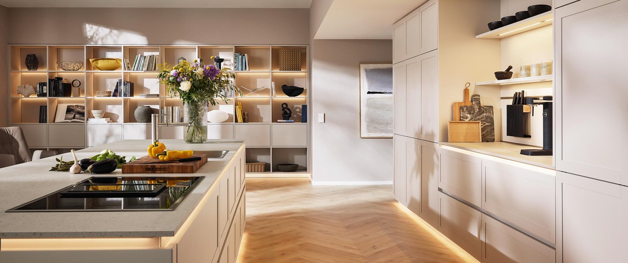 Eine moderne Küche mit hellen Holzschränken, einer Kochinsel, offenen Regalen mit Dekoartikeln, Unterbauleuchten, einer Vase mit Blumen und einem gemütlichen, minimalistischen Design in warmen, neutralen Farbtönen – sorgfältig gestaltet durch fachkundige Küchenplanung, um sowohl Funktionalität als auch Stil optimal zur Geltung zu bringen.